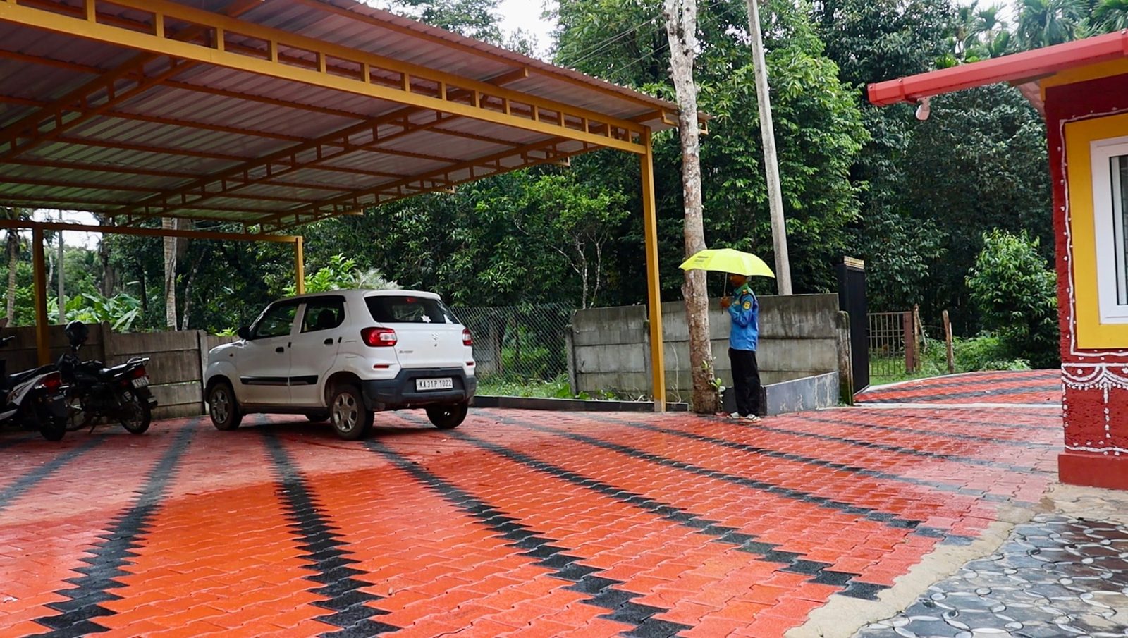 A covered parking area with a car and two-wheelers on red paved brick